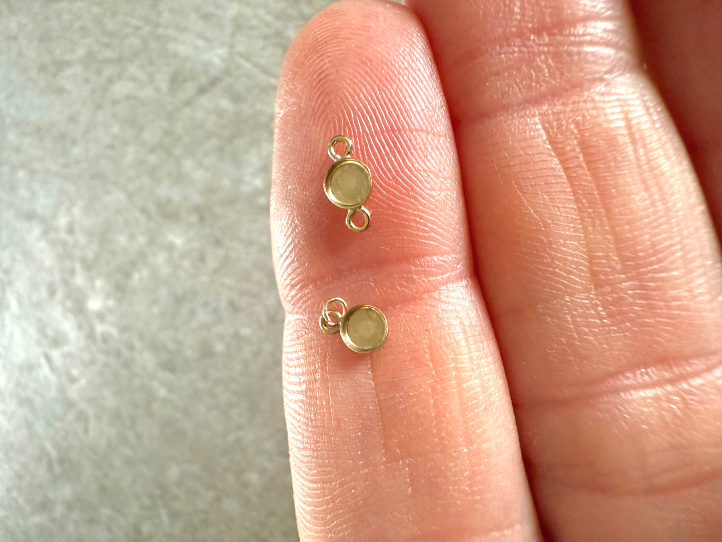 Two gold jewelry components on a person's finger against a neutral background